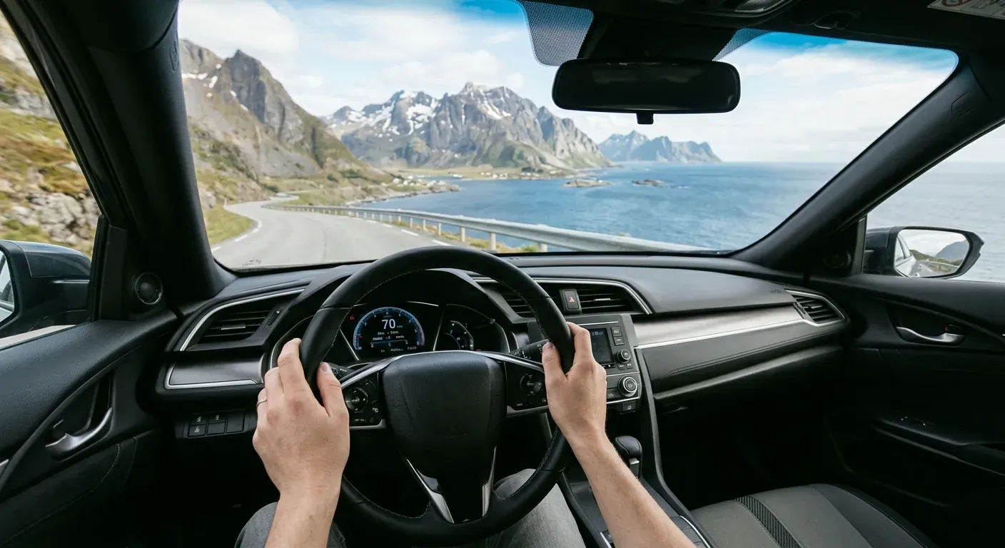 A driver's perspective from inside a modern car looking out onto a winding coastal road in Northern Norway, with steep mountains diving into the sea